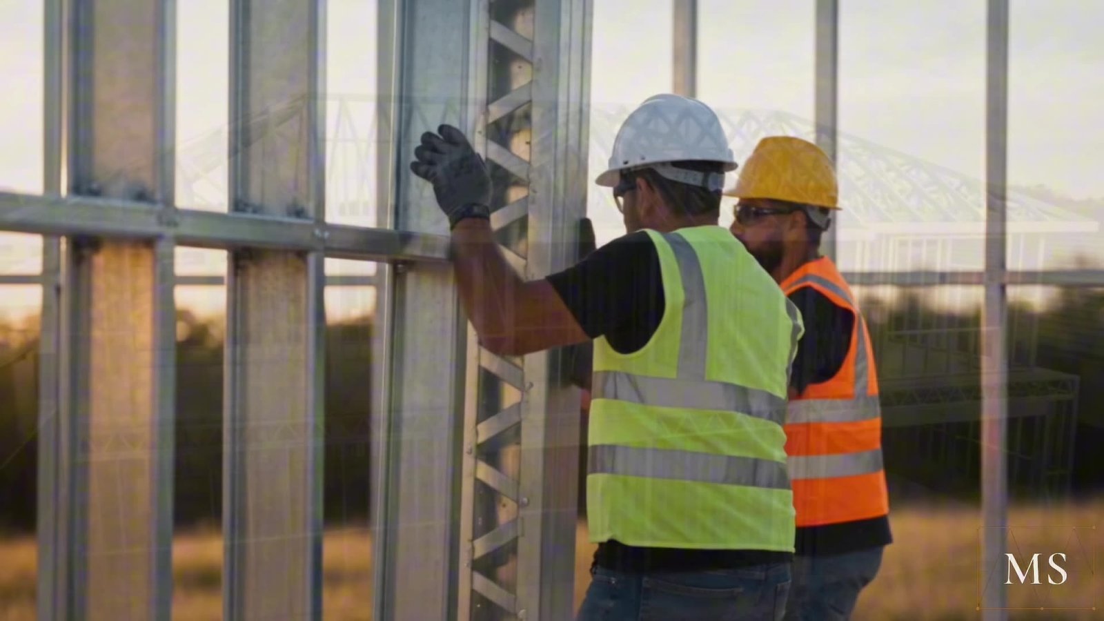 Dois montadores com capacete e colete posicionando um painel de perfis de aço galvanizado, ao entardecer.